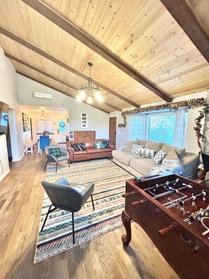 A spacious living area with a high ceiling, wooden beams, a sofa set, a patterned rug, and a foosball table.