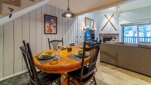 A dining area with a wooden table set for six, surrounded by chairs. There is a fireplace with a stone accent, a large window with a view of trees, and a wall-mounted mirror. - Chamonix 10: Charming, Walk to Canyon Lodge (Mammoth Lakes)