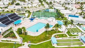 An aerial view of a resort with a swimming pool, tennis courts, and a large building with solar panels.