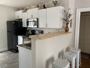 A compact kitchen with white cabinets, a black refrigerator, a microwave, and a stove. There are two white stools and a wooden countertop.