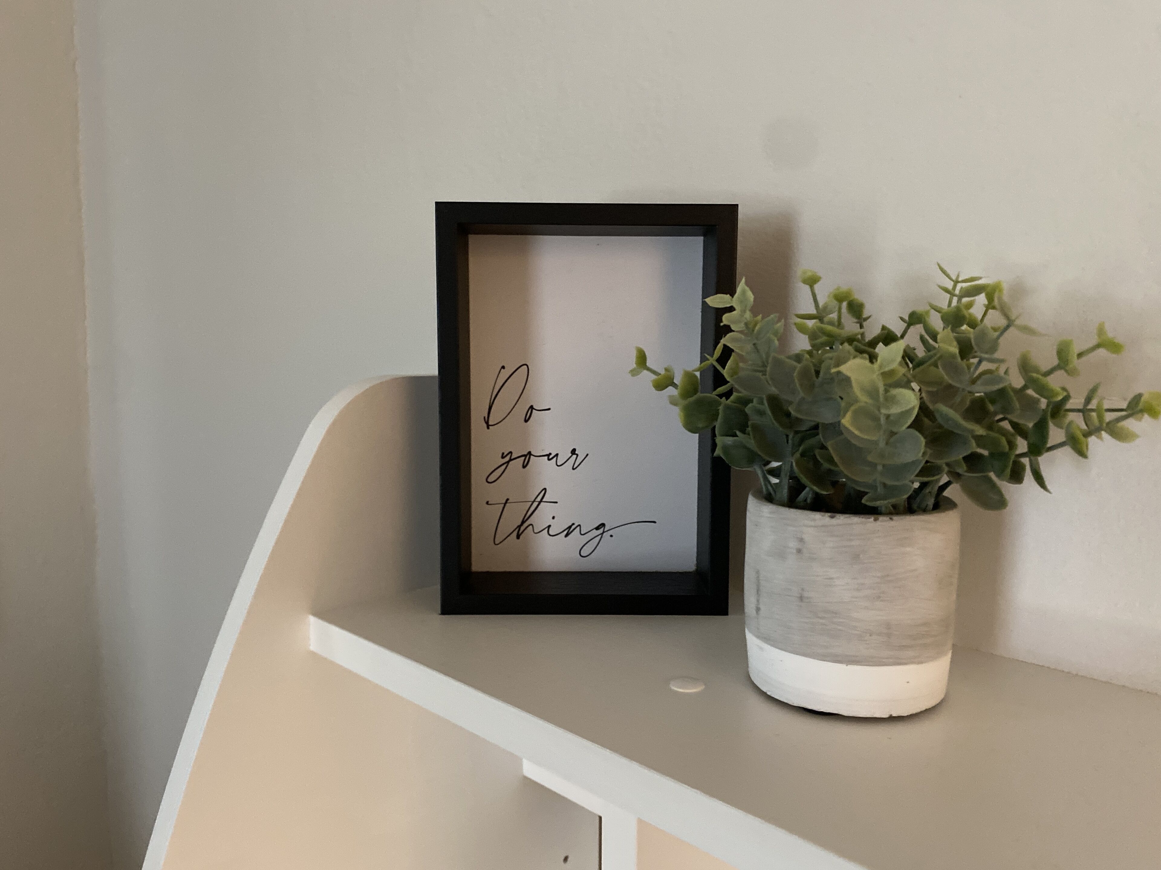 Un cadre contenant une citation inspirante accroché au mur, une plante en pot à côté, et une étagère blanche.
