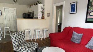 A living room with a red sofa, patterned armchair, and a small white table. There are three white stools and a kitchen area in the background.