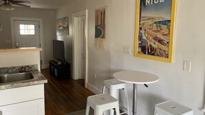 A compact kitchen with a sink, a ceiling fan, and a TV. There is a dining area with a table and two stools. A painting with the word "NICE" is on the wall.