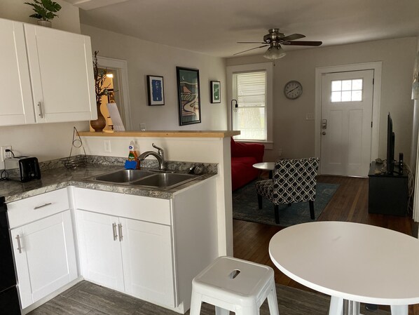 A compact kitchen with a sink, a small dining area with a white table and a patterned chair, a red couch, and a ceiling fan.