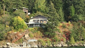 Et moderne hus med en stor terrasse med udsigt over en sø, omgivet af tæt skov og efterårsfarver.
