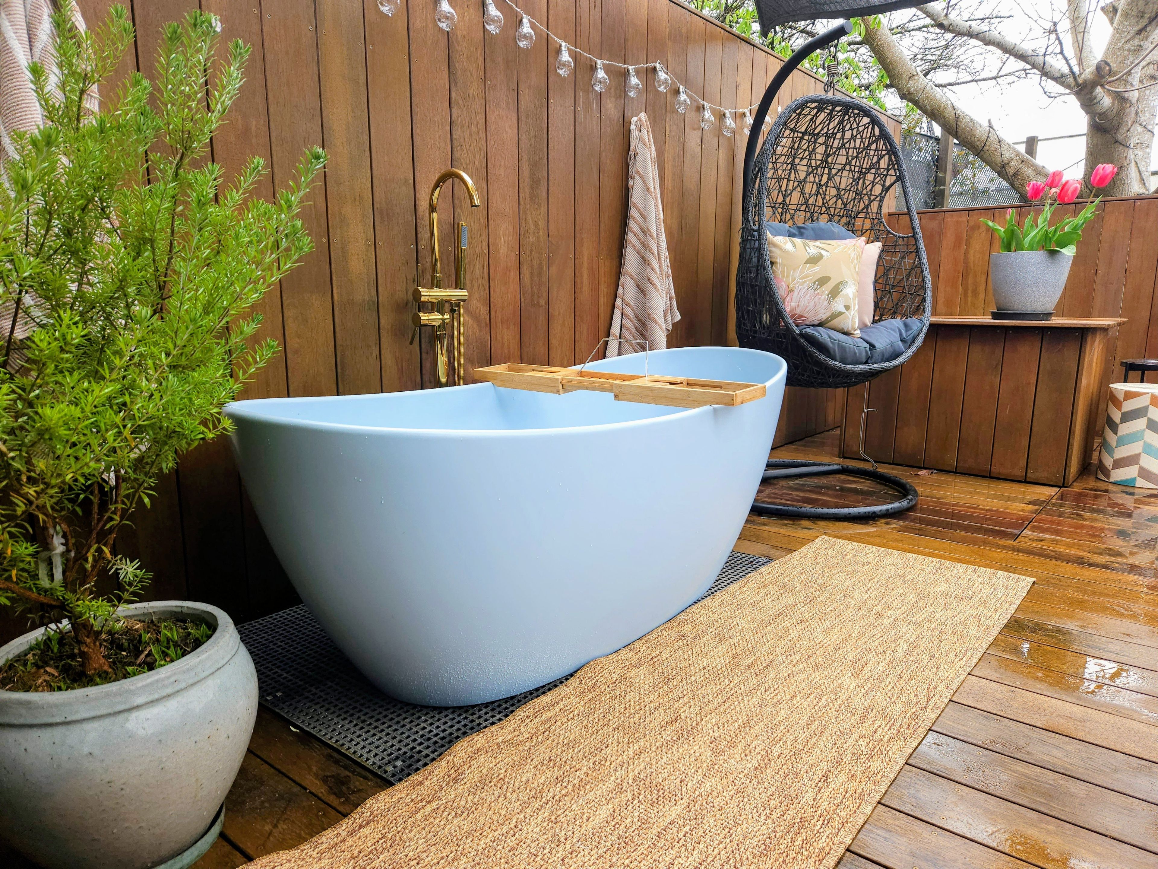 Een groot blauw bad met een houten zitje, een potplant, een rieten stoel en een hangende lichtslingering.