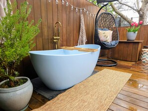 Una bañera azul grande con un asiento de madera, una planta en maceta, un sillón de mimbre y una guirnalda de luces colgantes.