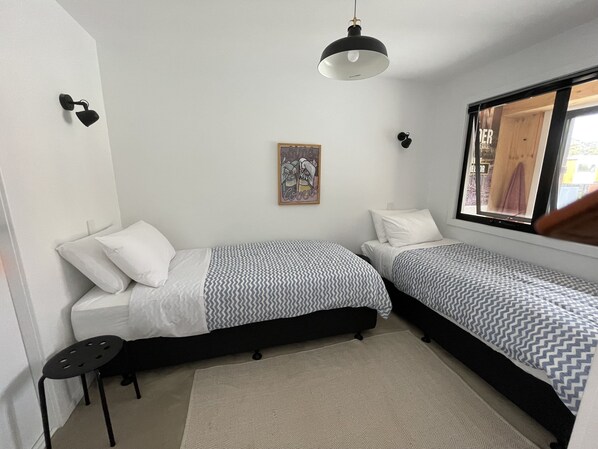 A small, modern bedroom with two single beds, a black chair, a patterned rug, a wall-mounted light, and a window with a view.