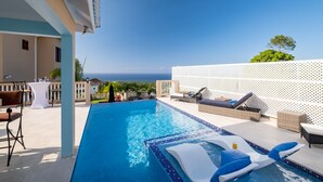 A swimming pool with lounge chairs and a table, overlooking a scenic view of the sea.