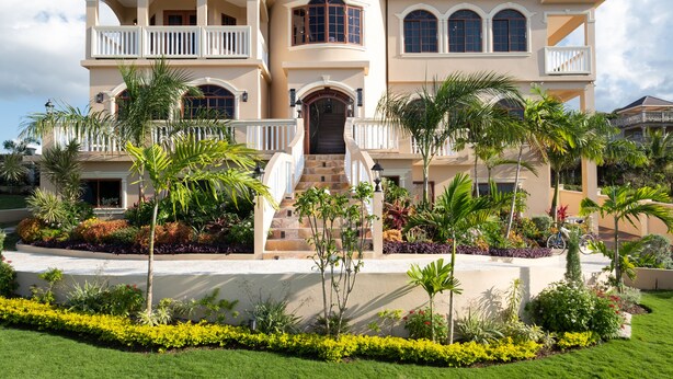 A large, two-story house with a balcony, a staircase leading to the entrance, and a well-maintained garden with various plants and a lawn.