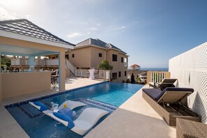 A swimming pool with lounge chairs and a hot tub, surrounded by a covered patio area and a large house with a balcony. - Luxury Villa · Ocean Views · Infinity Pool · Private Chef & Butler · Sleeps 8–10 (Discovery Bay)
