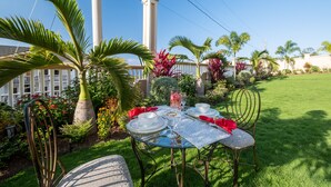 An outdoor dining area with a table set for two, surrounded by lush greenery and a white fence.