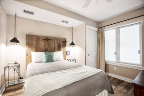 Een slaapkamer met een houten hoofdeinde, twee bedlampjes, een bed met een wit en grijs dekbedovertrek, een klein nachtkastje met een lamp, en een raam met zonwering.
