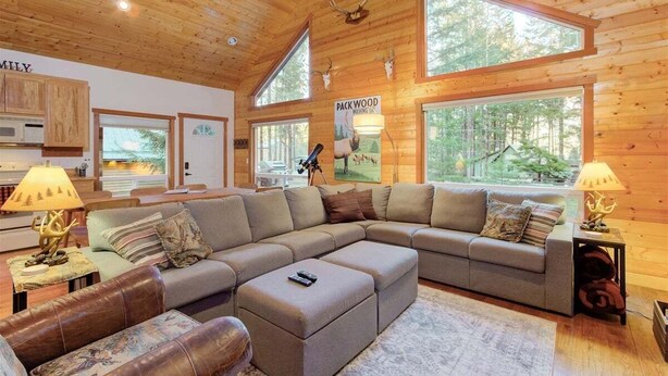 Interior acogedor de una cabaña con un amplio sofá modular en forma de L, una zona de comedor y grandes ventanales con vistas al bosque.