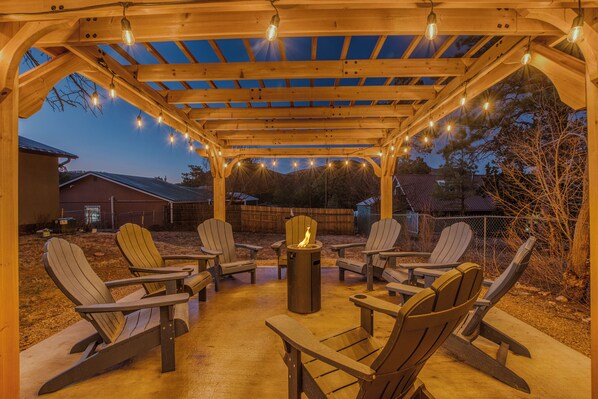 A wooden pergola with string lights and Adirondack chairs arranged around a central fire pit.