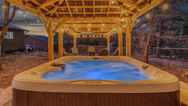 A hot tub under a wooden pergola with string lights, surrounded by chairs and a fence.