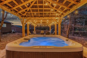A hot tub under a wooden pergola with string lights, surrounded by chairs and a fence.