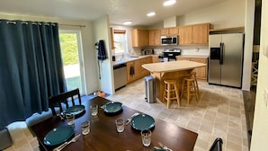 A modern kitchen with wooden cabinets, a central island, and a dining area with a table set for four. - Little Woods Outpost | Wifi, AC, Hot tub (Randle)