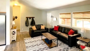 A living room with a sectional sofa, a coffee table, and a patterned rug. There is a coat rack with jackets and a door with a coat hanging on it. - Little Woods Outpost | Wifi, AC, Hot tub (Randle)
