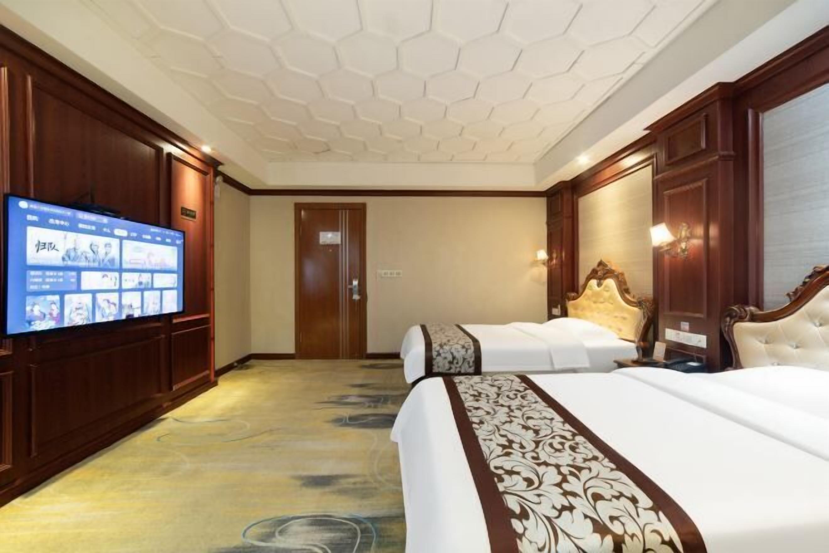 A hotel room with a large flat-screen TV, two beds, a wooden headboard, and a patterned carpet.