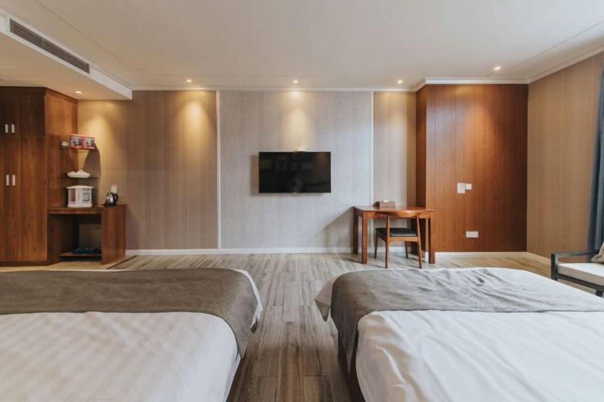 Habitación de hotel con dos camas, televisor montado en la pared, un escritorio pequeño y paneles de madera.