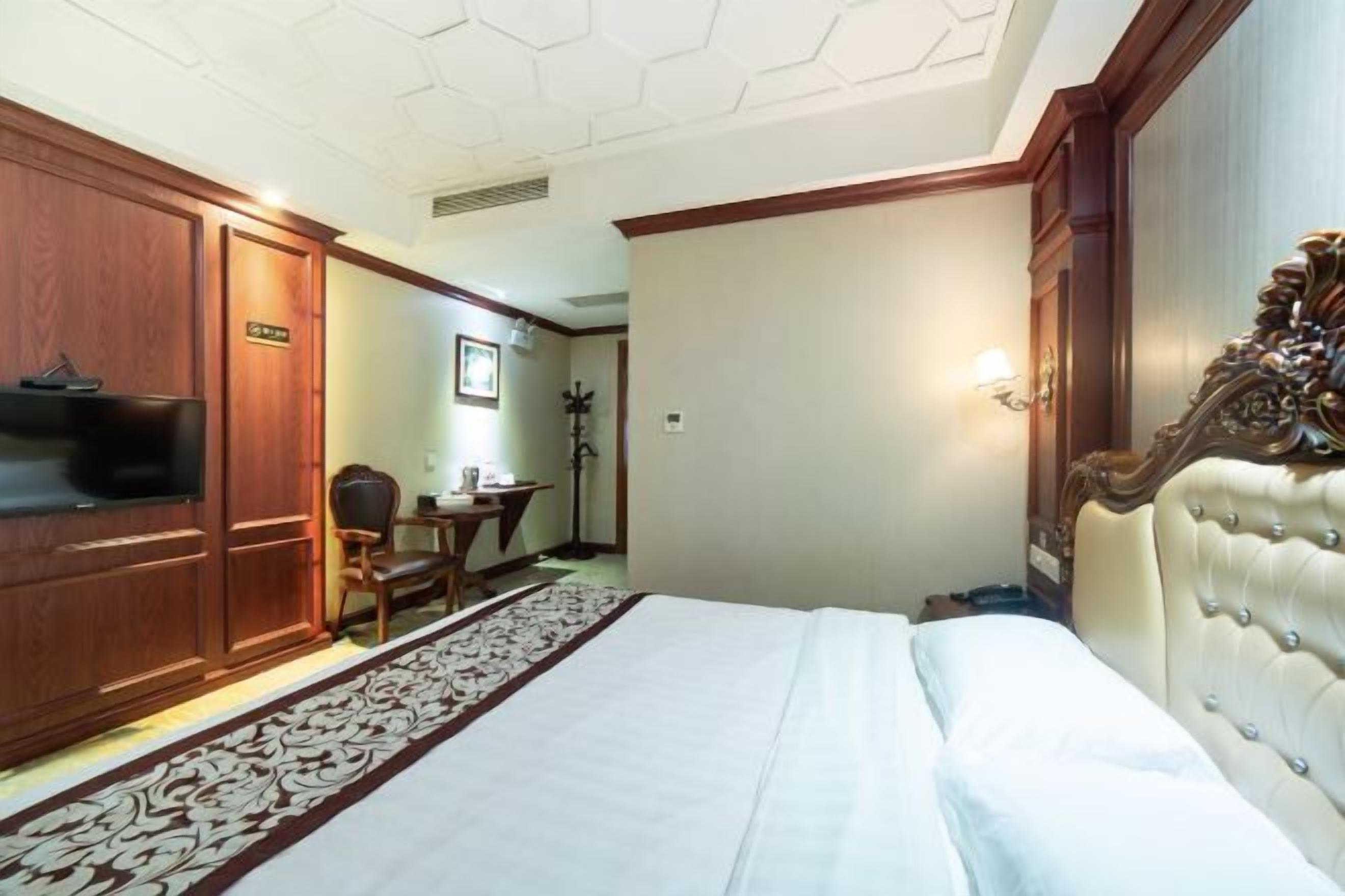 A hotel room with a bed, wooden wardrobe, television, desk, chair, and a decorative headboard.