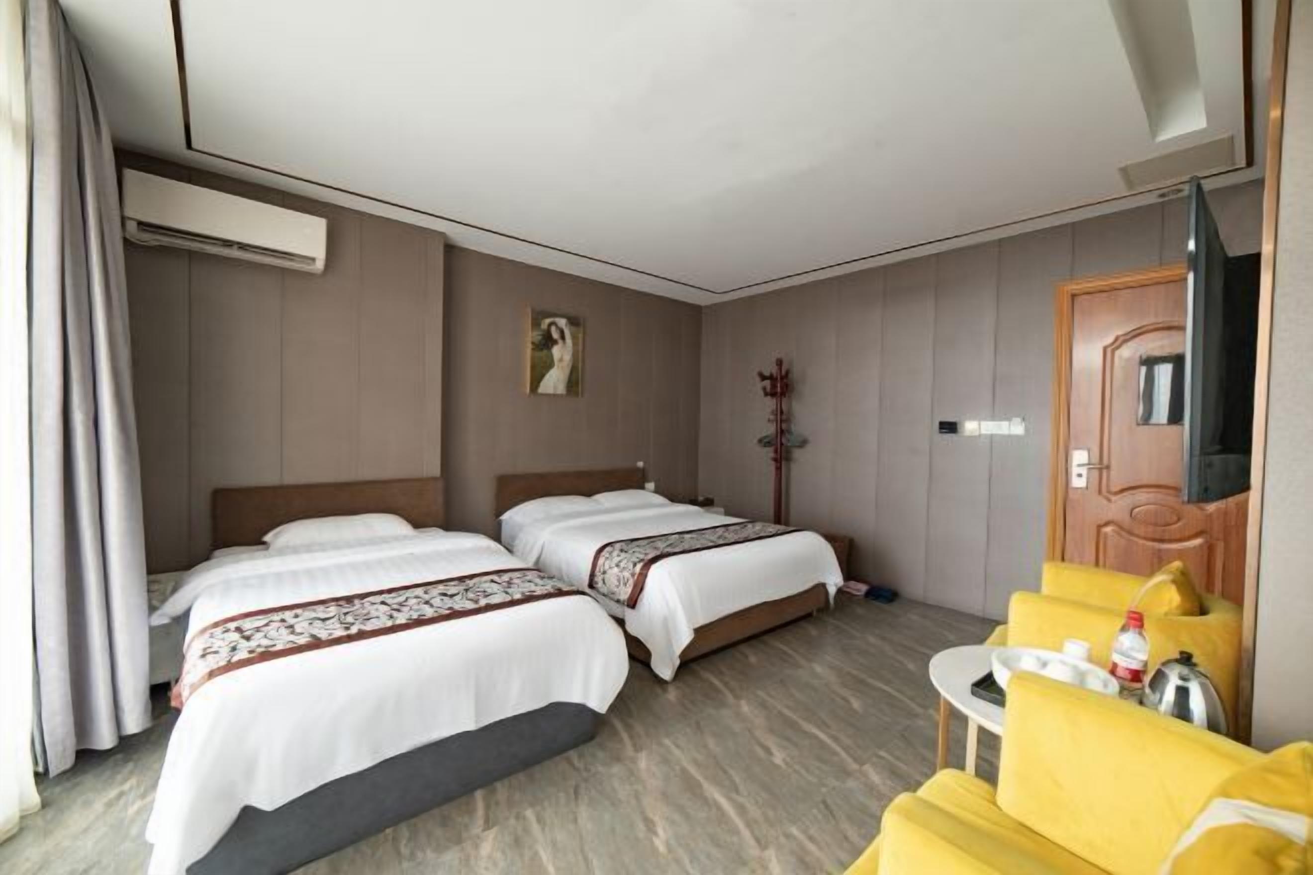 A hotel room with two beds, a small table with a kettle, a yellow armchair, and a wooden door.
