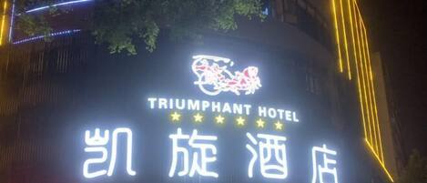 The exterior of the Triumphant Hotel with its illuminated signage and a visible entrance.