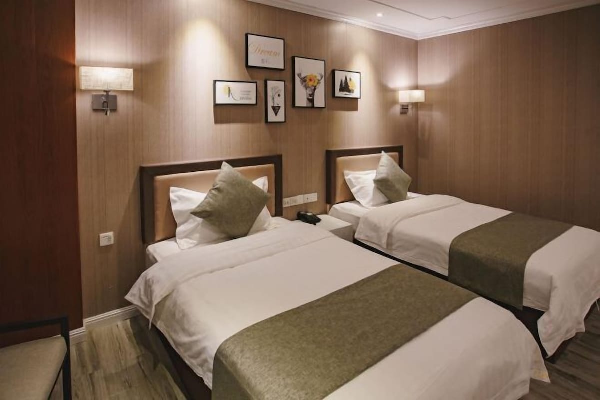 A hotel room with two beds, each with a headboard, white bedding, and a green throw blanket. There are wall-mounted lamps and framed pictures on the wall.