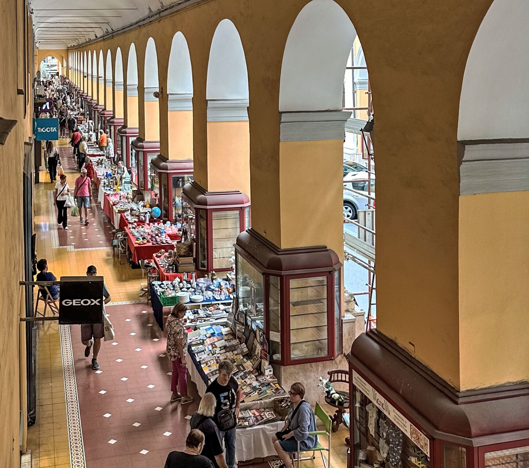 An indoor market with multiple stalls selling various goods, a GEOX sign, and a tiled floor.