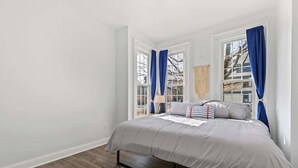 A bedroom with a large bed, two windows with blue curtains, a wooden floor, and a view of buildings outside. - Casa Soleada in Heart of East Rock 3BR | Cozy King Bed | Near DT/Yale (New Haven)