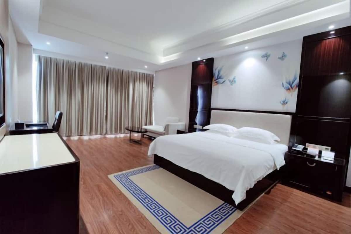 A modern hotel room with a large bed, a desk, and a chair. There is a large window with curtains, a wall-mounted TV, and a decorative wall piece.