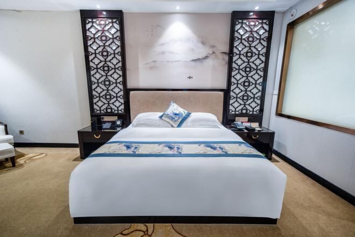 A hotel room with a large bed, two nightstands, a decorative wall panel, and a large window.