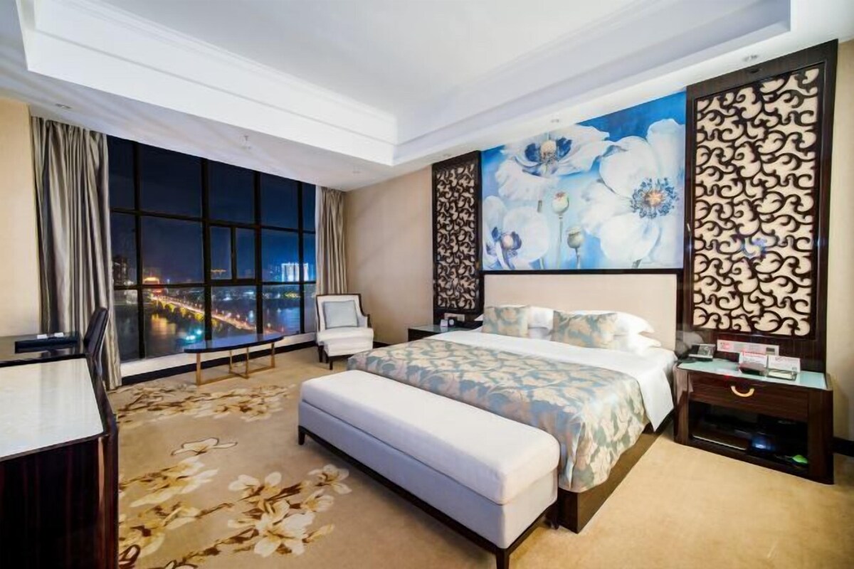 A hotel room with a large window, a bed with a floral bedspread, a bench, a desk, and a decorative wall panel.