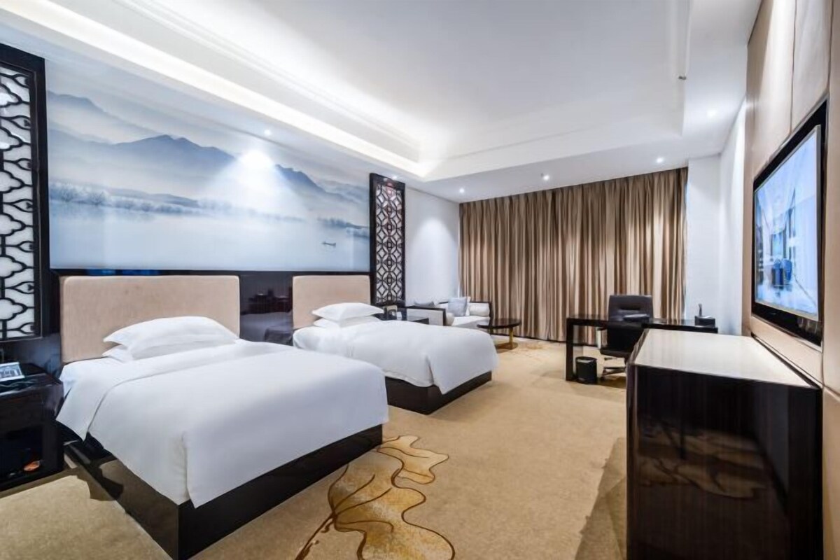 A hotel room with two beds, a TV, a desk, and a large window.