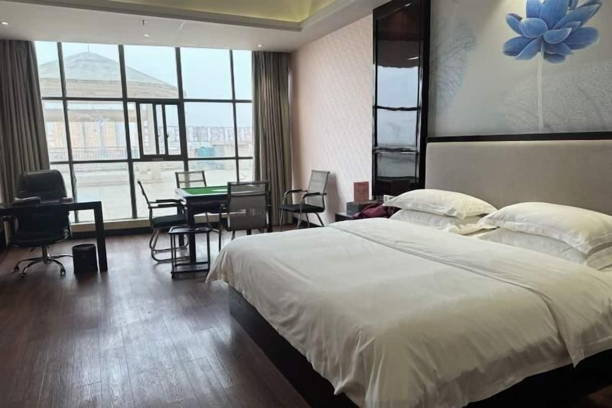 A hotel room with a large bed, a desk with a chair, a small table, and a view of a building through large windows.