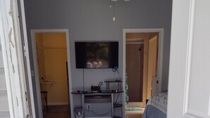 Una stanza con ventilatore a soffitto, TV a schermo piatto montata a parete, un tavolino con lampada, un letto con copripiumino decorato e un tappeto sul pavimento.