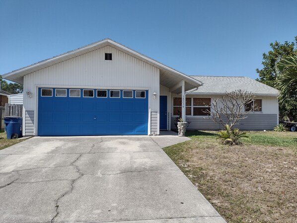 A single-story house with a blue garage door, a small porch, and a front yard with a tree and a planter.