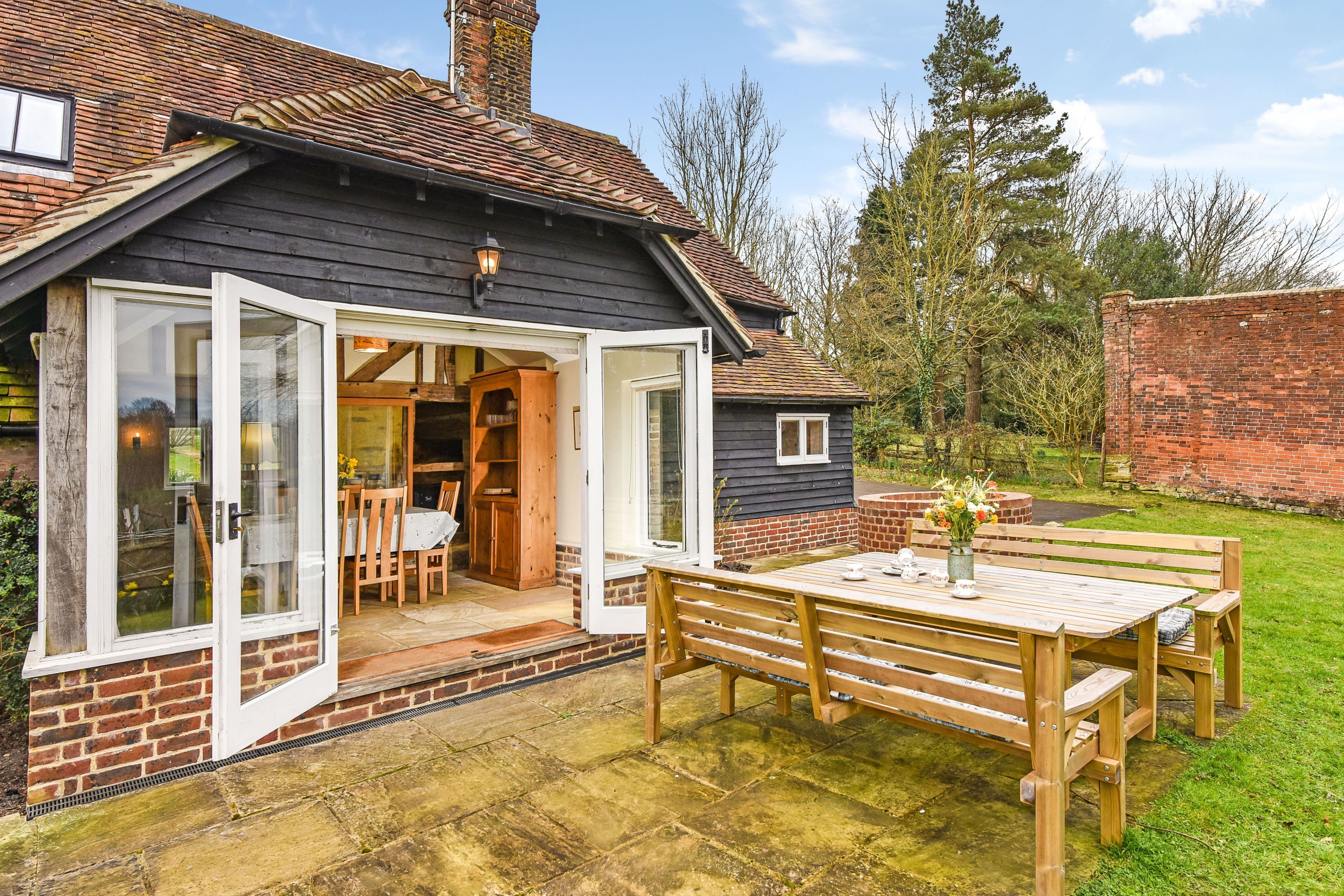 A house with a patio area featuring a wooden table and benches, a brick exterior, and a glass door leading to an interior space with a dining table and chairs.