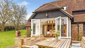 A wooden deck with a table and benches, a brick wall, and a glass door leading inside. - Bellflower Cottage - Ashdown Forest (Haywards Heath)