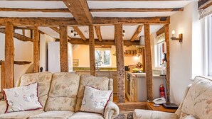 A rustic interior with exposed wooden beams, a sofa with floral upholstery, and a kitchen in the background. - Bellflower Cottage - Ashdown Forest (Haywards Heath)