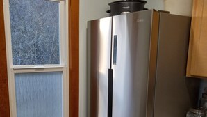 A kitchen with a stainless steel refrigerator, a window with a view of snow-covered trees, and a wooden floor.
