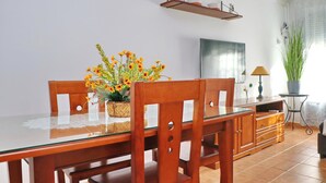 A dining area with a glass table, wooden chairs, a woven basket with flowers, a shelf with cups, and a potted plant.
