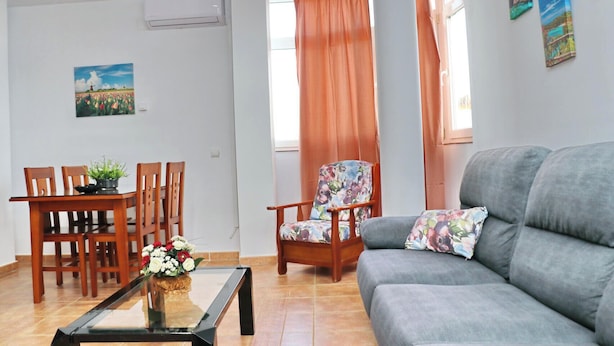 A living room with a grey sofa, wooden dining table with chairs, a floral armchair, and a glass coffee table with a flower vase.