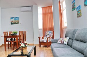 A living room with a grey sofa, wooden dining table with chairs, a floral armchair, and a glass coffee table with a flower vase.