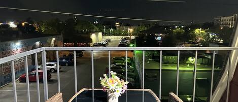 Un balcon avec des fauteuils en osier et une table, d’où l’on a vue sur un parking et des immeubles la nuit.