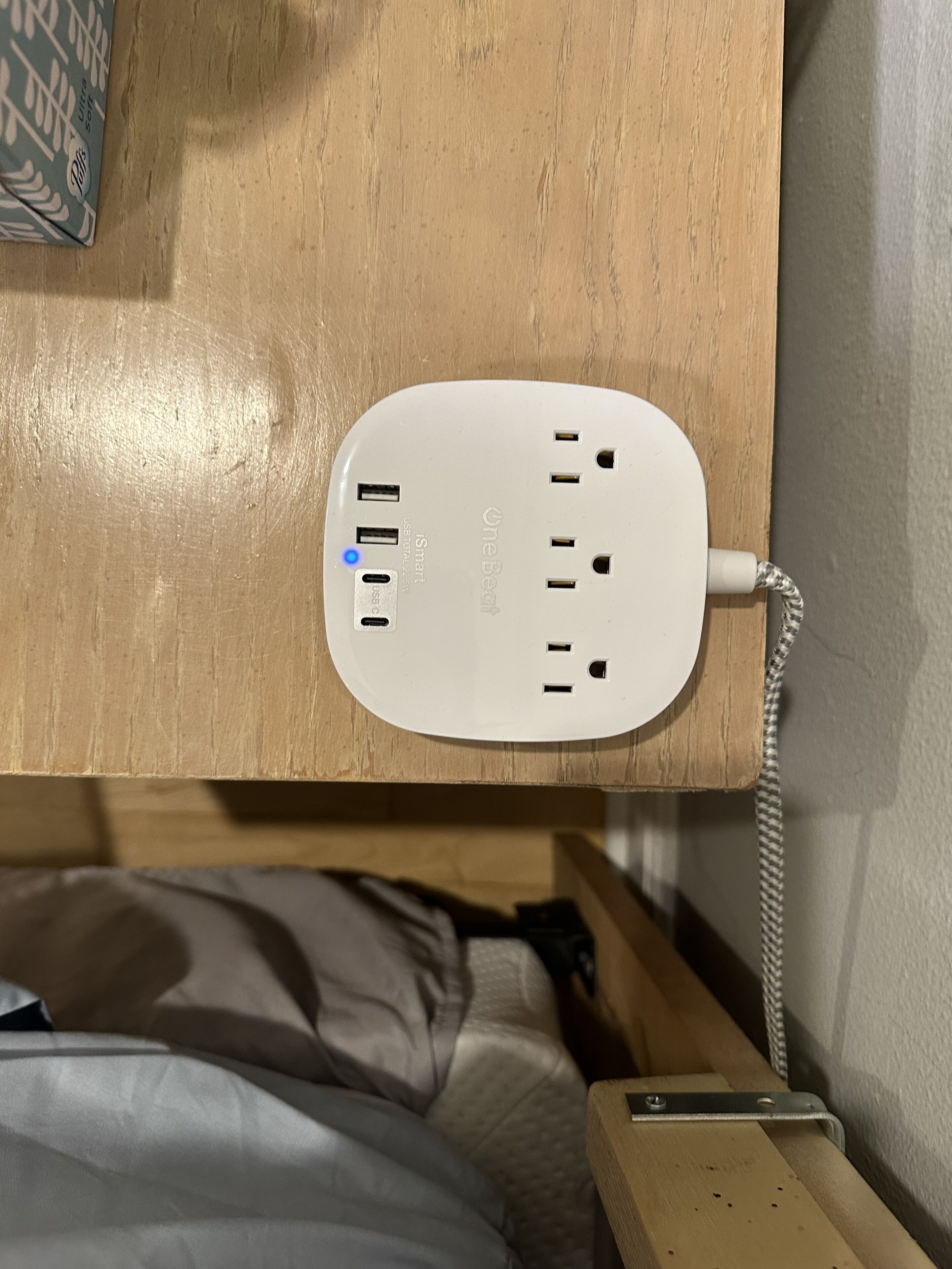 A white power strip with multiple USB ports and a power outlet, mounted on a wooden surface above a bed.