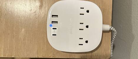 A white power strip with multiple USB ports and a power outlet, mounted on a wooden surface above a bed.