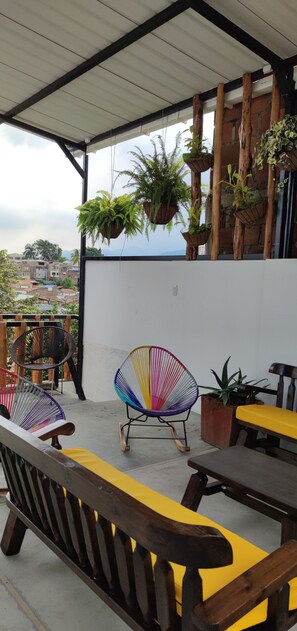 Un espace extérieur abrité, doté de bancs en bois et de chaises colorées, de plantes en pot, et offrant une vue sur le paysage urbain.