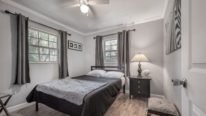 Une chambre à coucher comprenant un lit, une table de chevet, un banc, un ventilateur de plafond et deux fenêtres avec des rideaux.